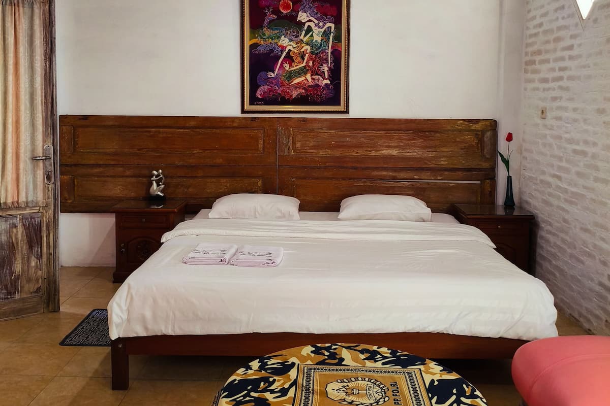 Quarto com cabeceira de madeira, cama com roupa de cama branca, um quadro colorido acima da cama, um criado-mudo pequeno, um banco de madeira com capa estampada e uma cadeira vermelha.