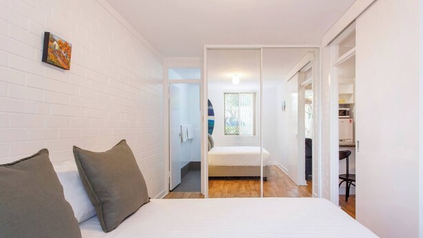 A bedroom with a bed, two pillows, a white brick wall, a small painting, and a view into the kitchen.