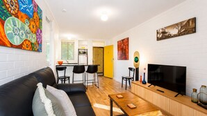 A modern living room with a black leather sofa, a wooden coffee table, a TV, and a kitchen area with a yellow door.