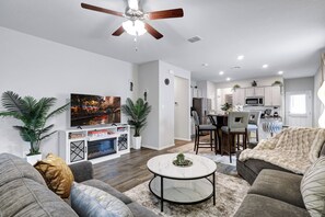 A modern living room with a sofa, coffee table, and a TV. There's a dining area with chairs and a kitchen in the background.