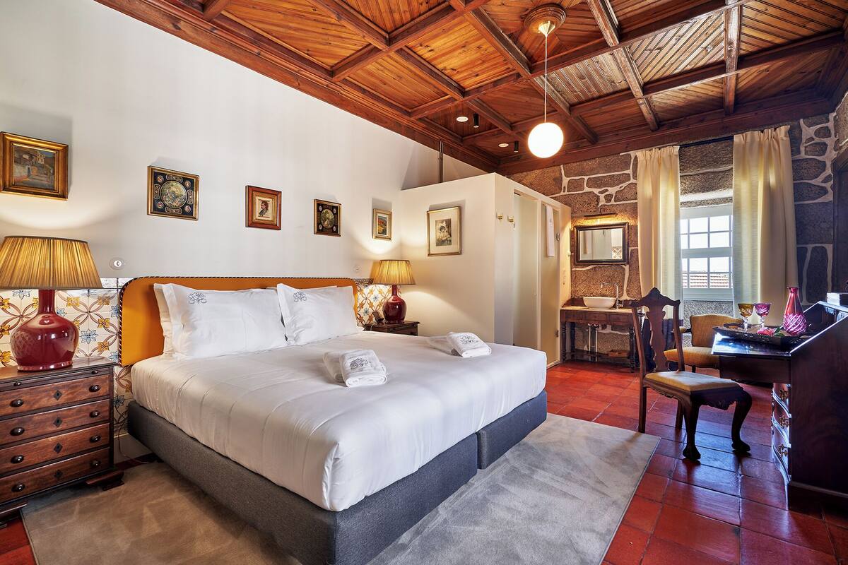 A bedroom with a large bed, wooden ceiling, stone walls, a desk, and a chair.