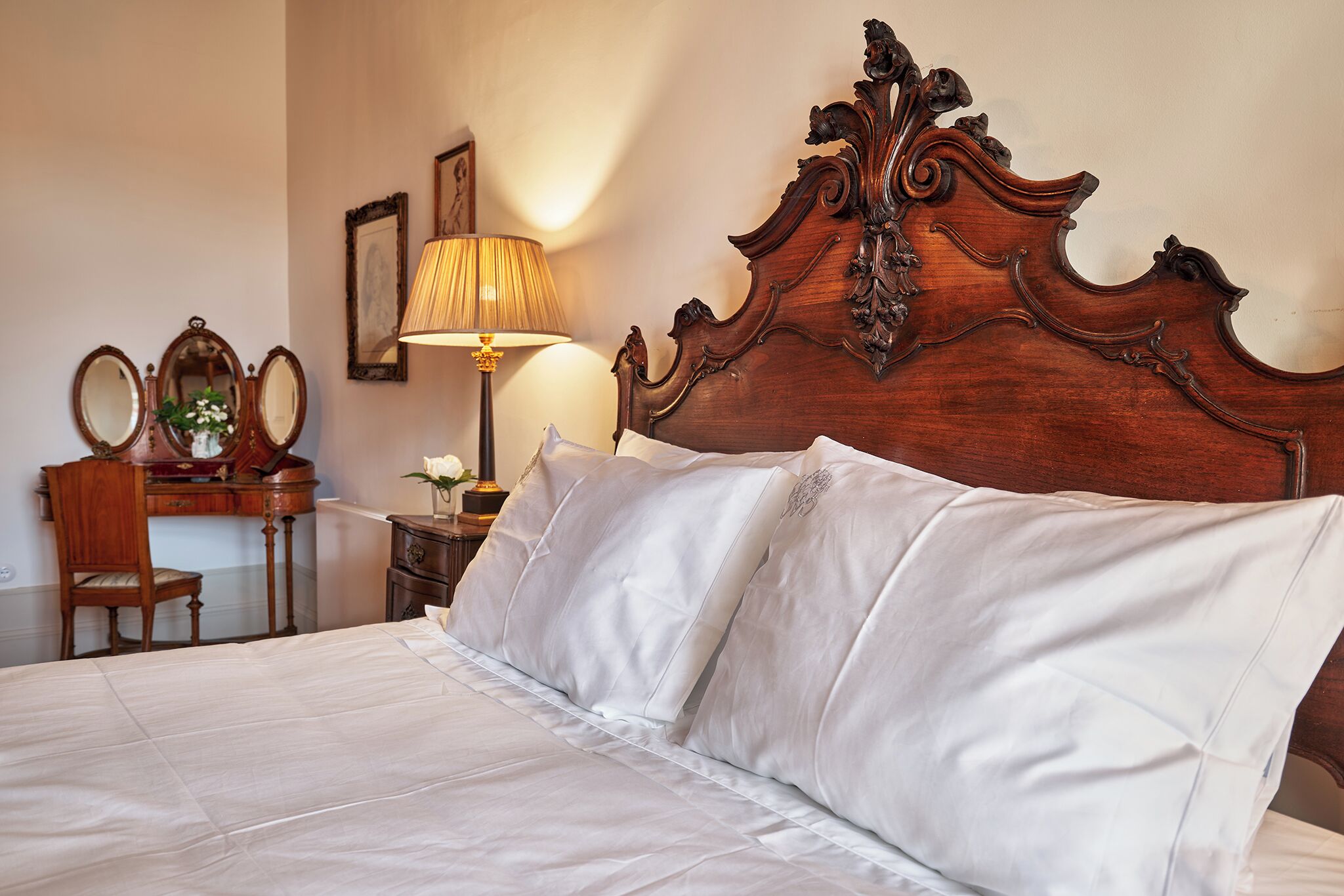 A neatly made bed with white linens, a wooden headboard with intricate carvings, a bedside table with a lamp, and a mirror on the wall.