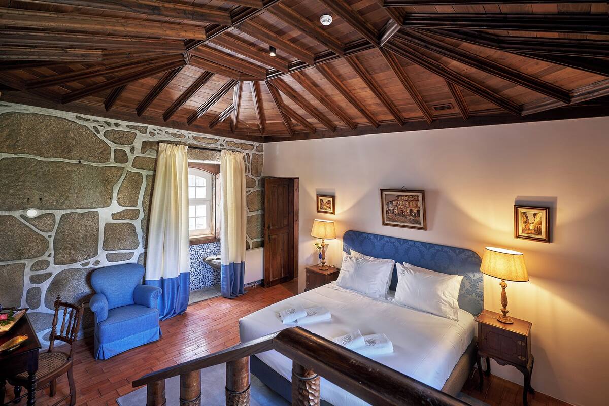 A bedroom with a stone wall, a wooden ceiling, a bed with white linens, a blue armchair, and two bedside lamps.