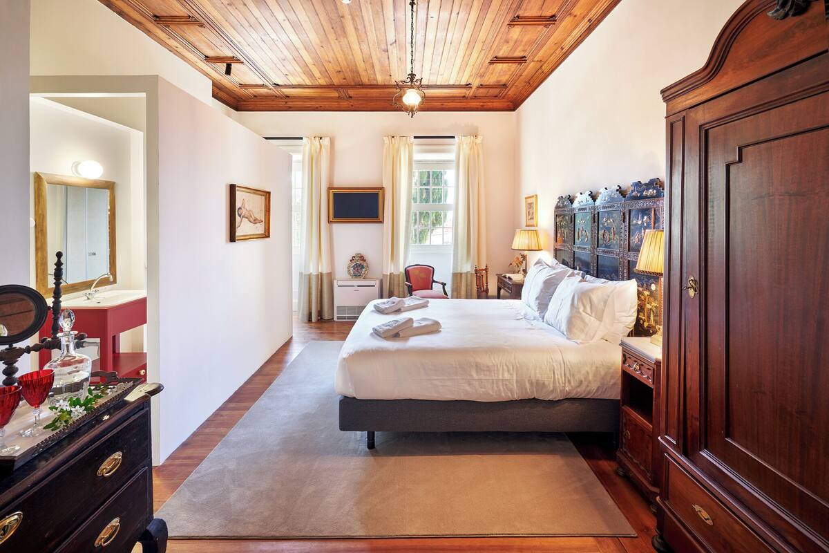 A hotel room with a wooden ceiling, a large bed, a dresser, a mirror, and a window with curtains.