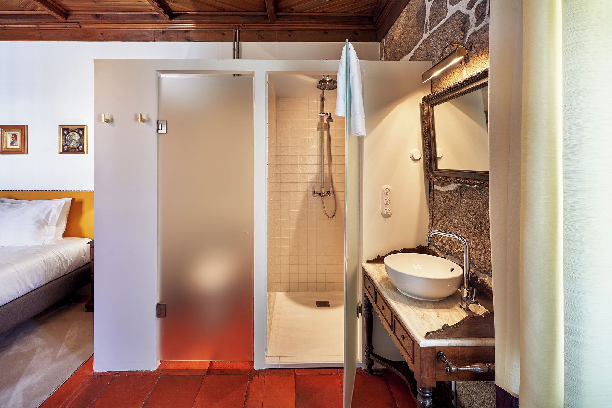 A bathroom with a shower, a sink, and a bed in the adjacent room.