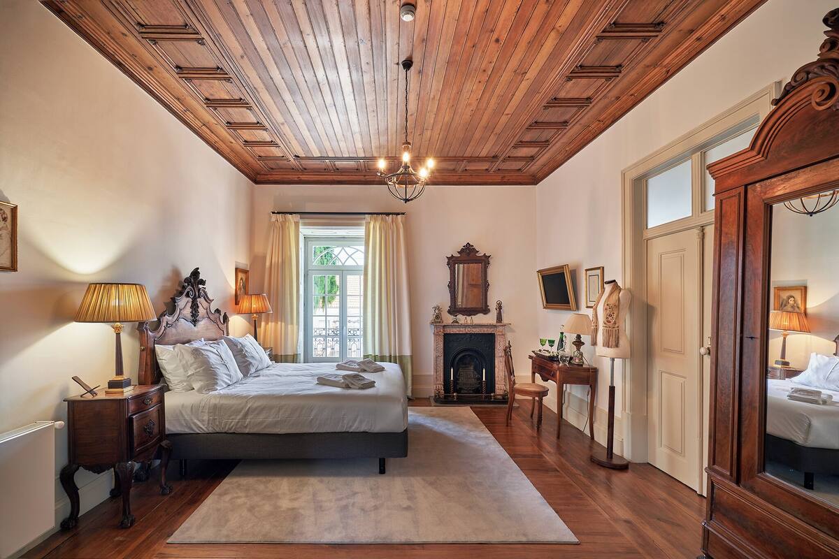 A bedroom with a wooden ceiling, a large bed, a fireplace, and a mirror.