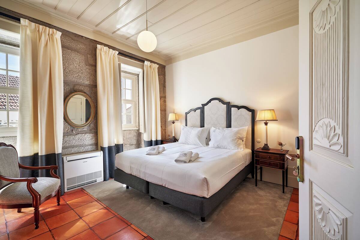 A bedroom with a large bed, a chair, a round mirror, and a nightstand with a lamp.