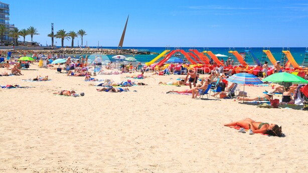 Una playa con gente tomando el sol, sombrillas coloridas y un tobogán de agua.