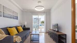Una sala de estar moderna con un sofá gris, cojines amarillos, una mesa de centro, un televisor sobre un mueble de madera y un balcón con vista a edificios y una piscina.