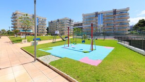 Un parque infantil con columpios y un tobogán, rodeado de un césped cuidado y un camino empedrado.