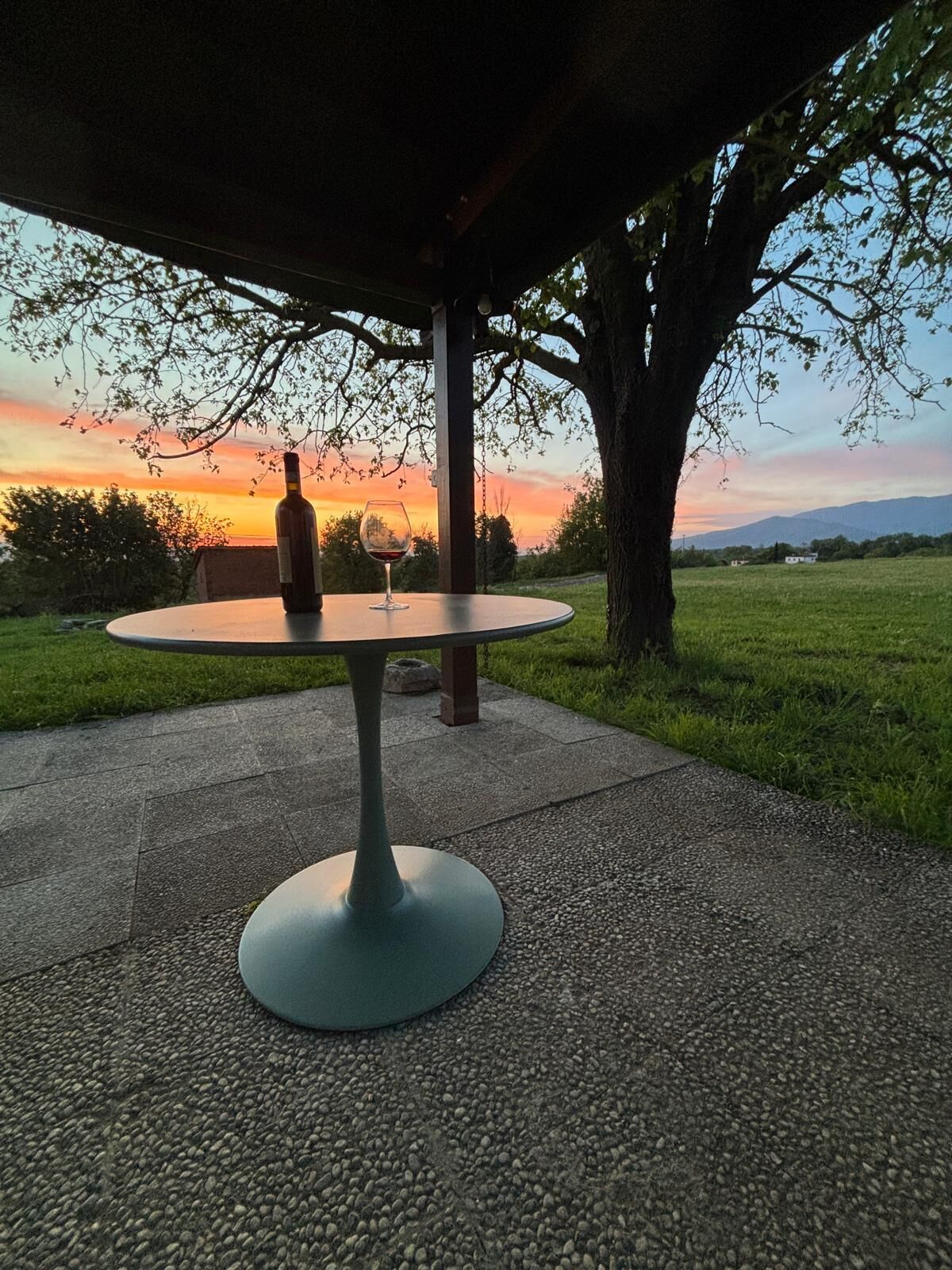 Et rundt bord med en flaske og et glas rødvin, placeret udendørs under en pergola med udsigt til en solnedgang og et træ.