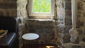 Una habitación rústica de piedra con techo de madera, una ventana pequeña que da a la vegetación, una mesa redonda, un sofá de cuero negro y una toalla blanca colgada en la pared.