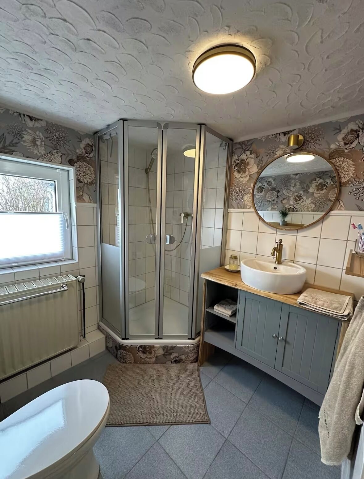 Une salle de bain avec une douche à parois de verre, un lavabo blanc, un miroir rond et une fenêtre.