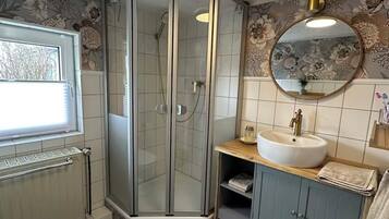 Une salle de bain avec une douche à parois de verre, un lavabo blanc, un miroir rond et une fenêtre.