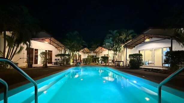 A nighttime view of a pool area with a clear pool, surrounded by buildings with large windows and palm trees. - vivi bungalows resort5 (Rawai)