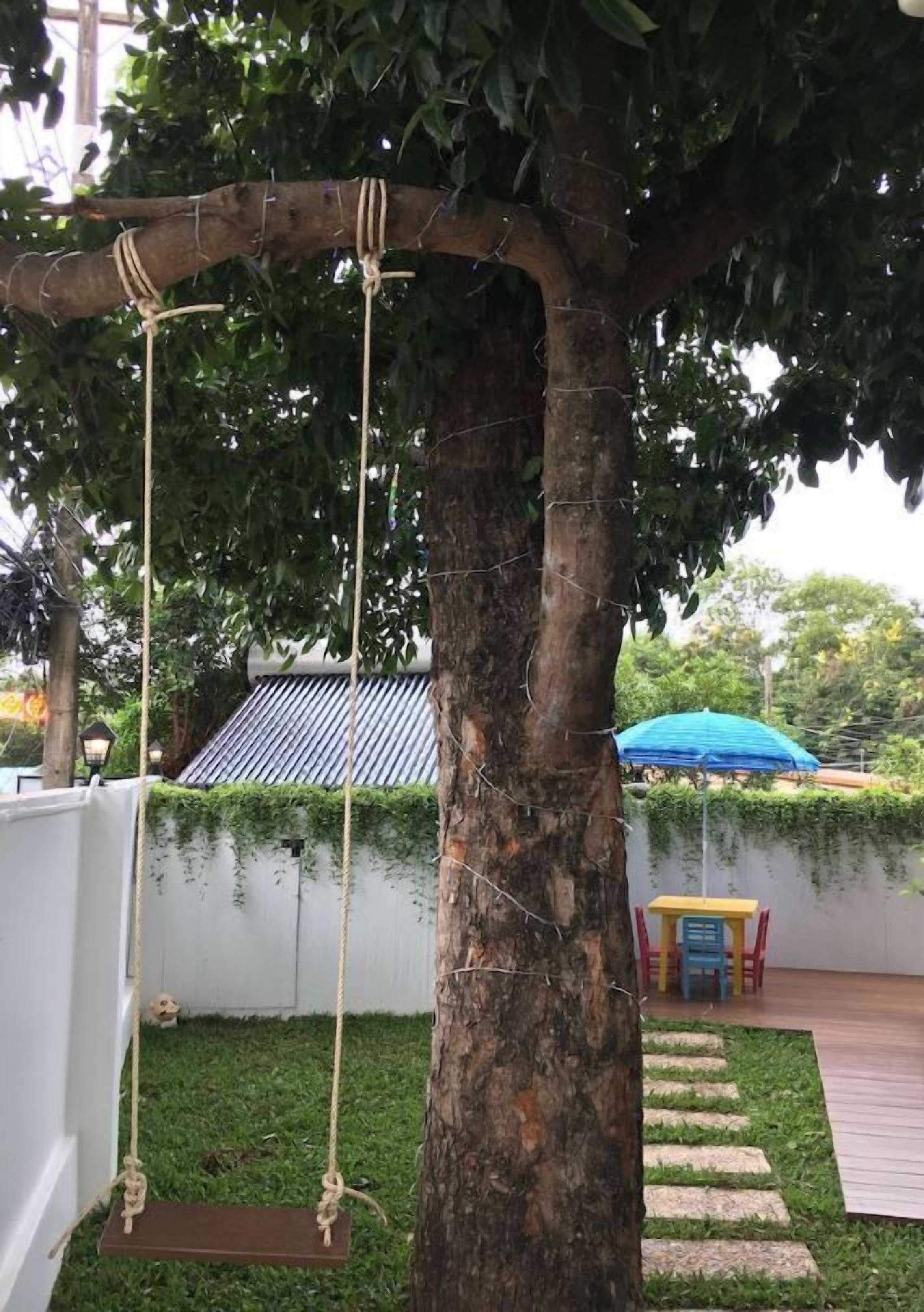 A tree swing attached to a tree, a small garden area with a stepping path, and a blue umbrella in the background.