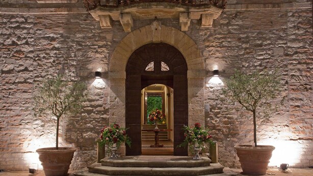 A stone building with a central arched doorway, flanked by two potted trees and illuminated by wall-mounted lights. - My Rental Homes - Villa dei Cappuccini with private pool, jacuzzi, air conditioning (Gubbio)
