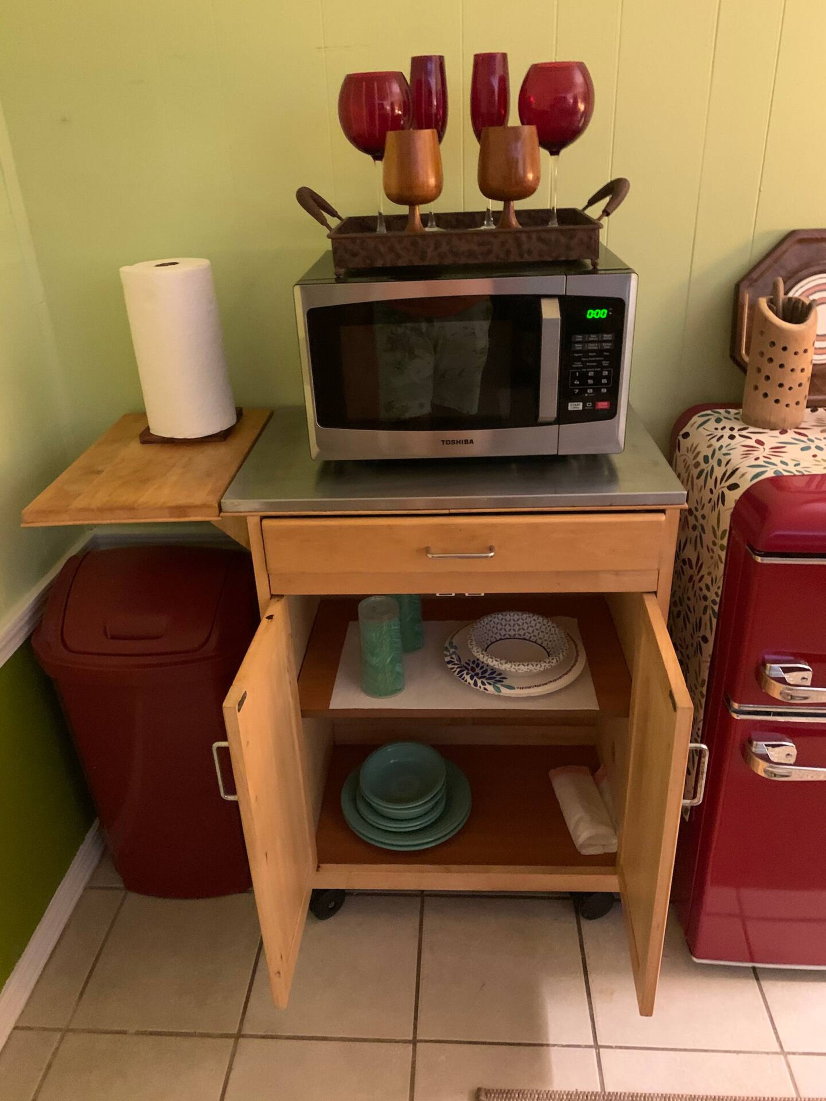Cocina compacta con microondas sobre una encimera de madera, un cajón pequeño con vajilla y un cubo de basura.