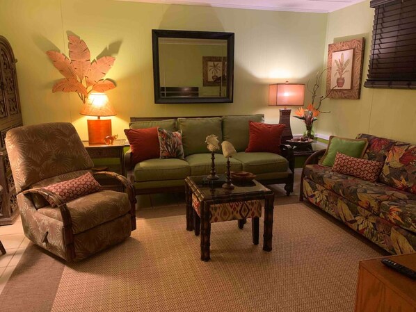 Una sala de estar acogedora con un sofá verde, un sillón marrón y un sofá estampado. Hay una mesa de centro de madera y una lámpara en una mesita auxiliar.