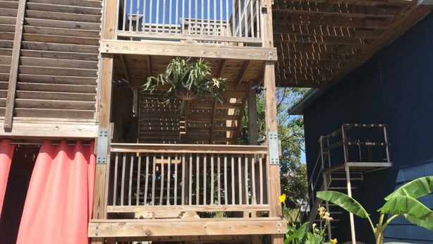 Eine mehrstöckige Holzkonstruktion mit Balkon, einem kleinen Gartenbereich mit Pflanzen und einer blauen Wand im Hintergrund.