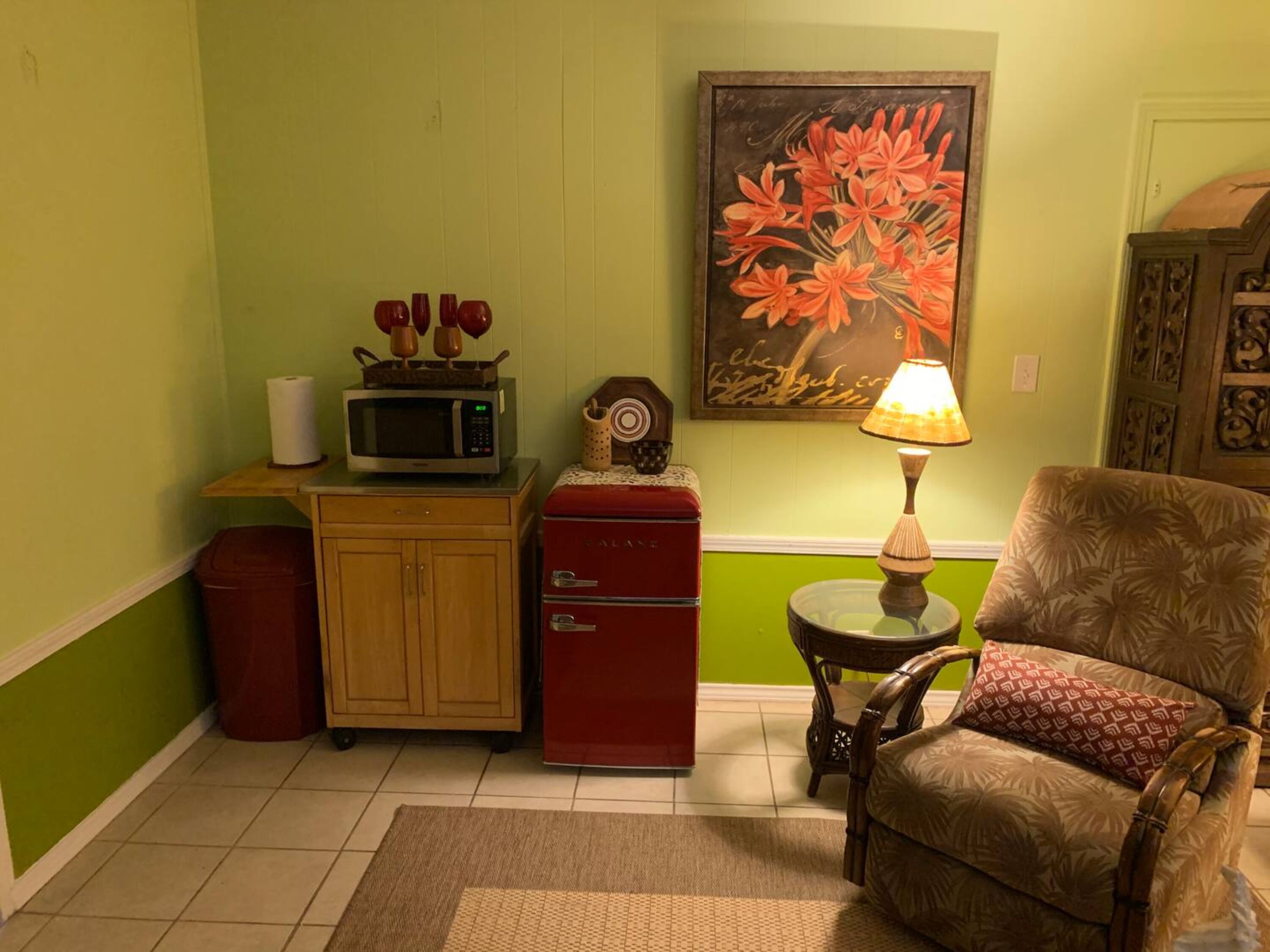 A compact living space with a microwave, a red refrigerator, a wooden cabinet, a chair, and a table with a lamp.