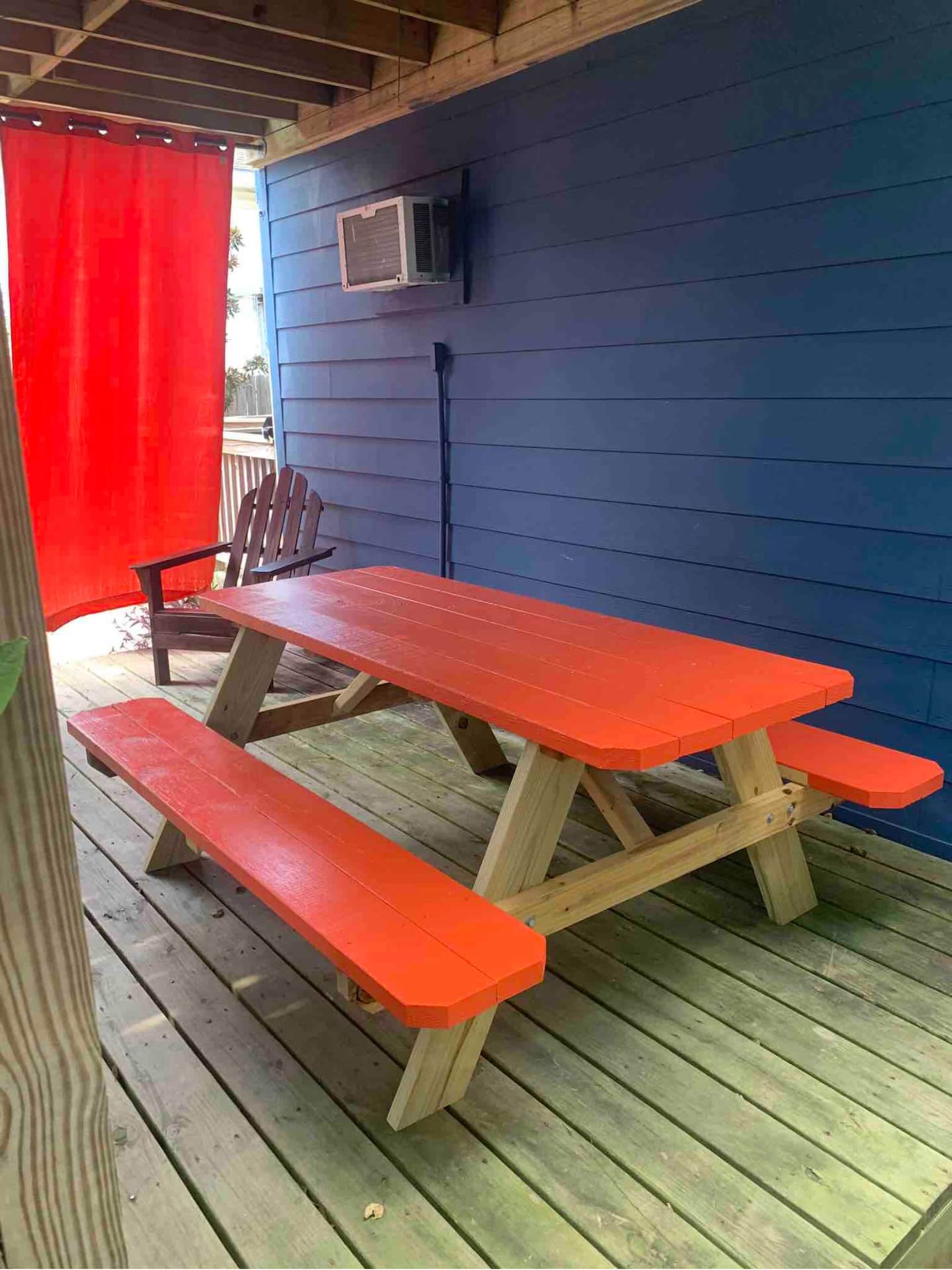 Una mesa de picnic de madera con bancos sobre un muelle de madera, una pared azul y un equipo de aire acondicionado.