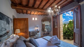 Ein Schlafzimmer mit einem großen Bett, einer Kristallleuchtre, einer Holzbalkendecke, einem Spiegel und Blick auf Grünflächen.