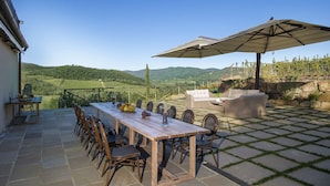 An outdoor patio with a wooden table and chairs, a wicker sofa, and a large umbrella. - Villa Serena with private pool - My Rental Homes (Greve In Chianti)