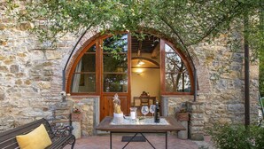 A stone building with a large arched window, a wooden table set with glasses and a candle, and a bench with cushions. - Villa Grevigiana pool and SPA - My Rental Homes (Greve In Chianti)