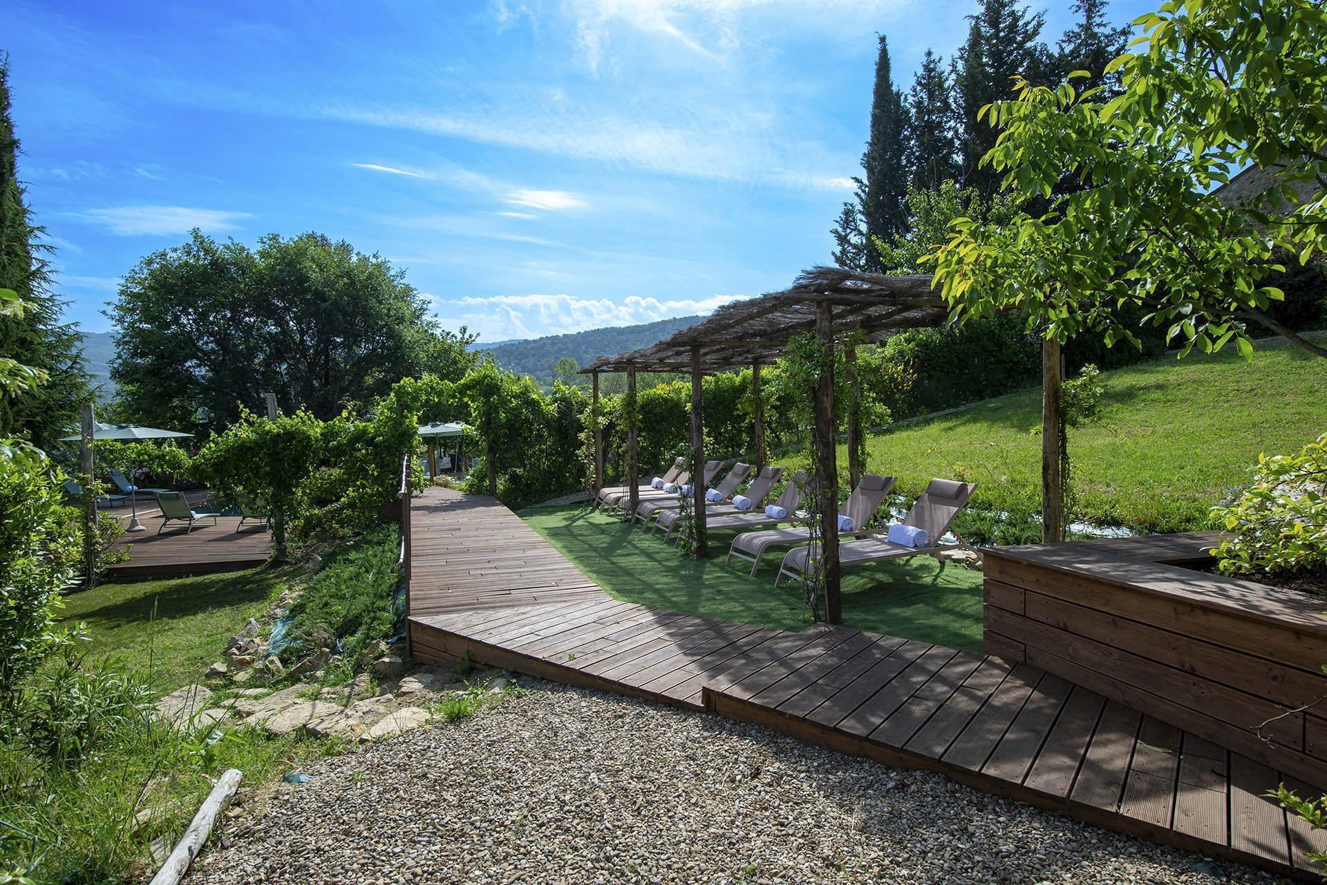 A wooden deck with lounge chairs and a pergola, surrounded by greenery and a view of hills.