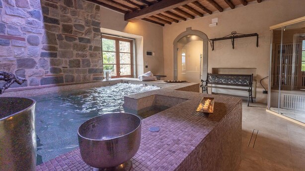 A spa area with a stone wall, a hot tub, a wooden bench, and a large metal bowl. - Villa Grevigiana pool and SPA - My Rental Homes (Greve In Chianti)