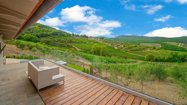 A balcony with a wicker sofa and a glass railing overlooking a vineyard. - Villa del fattore with pool - My Rental Homes (Greve In Chianti)
