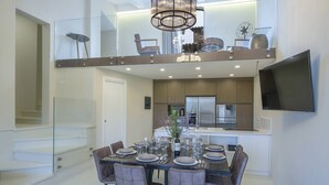 A modern dining area with a glass-top table set for six, surrounded by six chairs. A kitchen with a stainless steel refrigerator and microwave is visible in the background. - Villa del fattore with pool - My Rental Homes (Greve In Chianti)