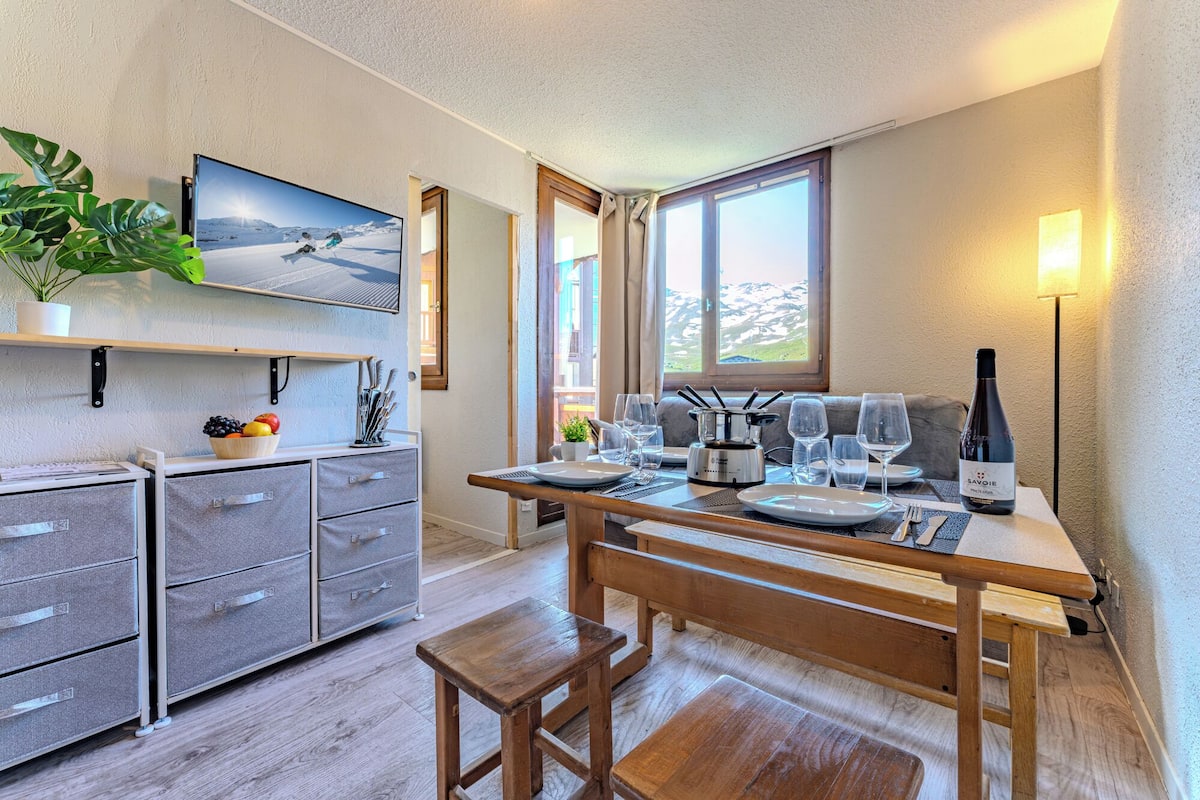 A modern kitchen with a wooden table set for dining, a TV mounted on the wall, and a bottle of wine on the table.