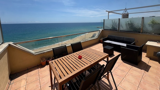 Eine Terrasse mit Holztisch und -stühlen, einer Glasreling und Blick aufs Meer.