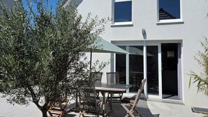 Una casa a due piani con un patio che include un tavolo, sedie, un piccolo albero e un'ampia porta scorrevole in vetro.