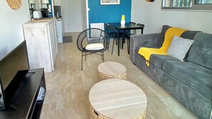 Een woonkamer met een grijze bank, een eettafel en een keuken met een koelkast en magnetron.