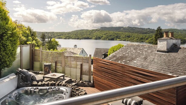 A rooftop patio with a hot tub, outdoor seating, and a scenic view of a lake and forested hills. - Poole Brae Lake View Getaway | Ridgestone (Bowness-on-Windermere)