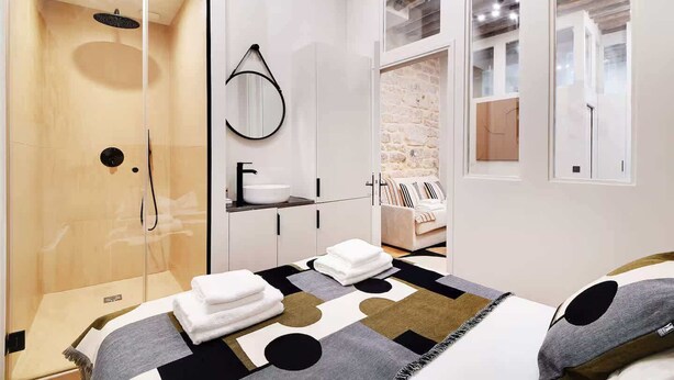 Ein modernes Schlafzimmer mit Glasdusche, großem Spiegel, Bett mit geometrischem Bettbezug und Steinwand.