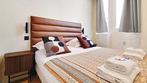 Una cama bien hecha con ropa de cama y almohadas estampadas, una mesita de noche de madera y una ventana con cortinas.