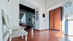 Un pasillo amplio con suelo de baldosas, una silla blanca, una puerta de madera y un reloj en la pared.
