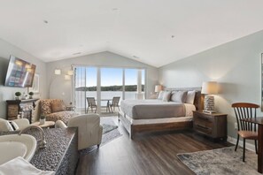 A spacious bedroom with a large bed, a seating area with a sofa and chairs, a small table, and a view of the outdoors through large windows.