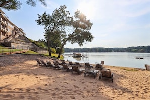 Une plage avec des chaises longues, un grand arbre et un bâtiment doté de balcons donnant sur un lac.