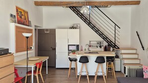 A compact kitchen with a central island, a dining area with a table and chairs, a staircase, and a living area with a sofa and decorative pillows.