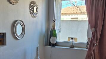 Une petite pièce bien éclairée, avec un bureau blanc, une chaise, une fenêtre donnant sur l’extérieur et une bouteille de vin posée sur le rebord de la fenêtre.