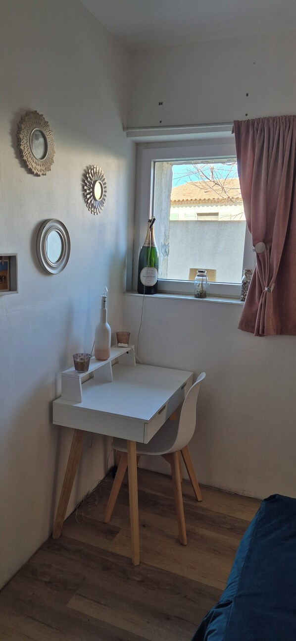 A small, well-lit room with a white desk, a chair, a window with a view of the outside, and a bottle of wine on the windowsill.