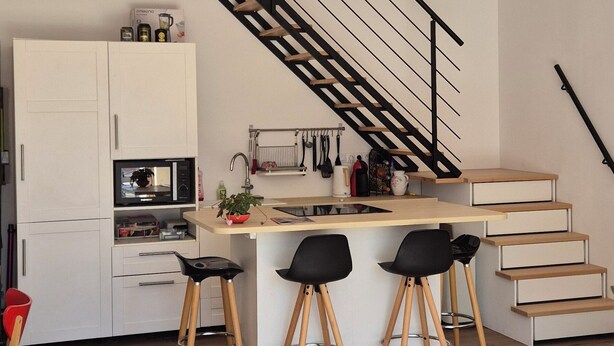 A modern kitchen with a central island, wooden beams, a staircase, and a dining area with stools.