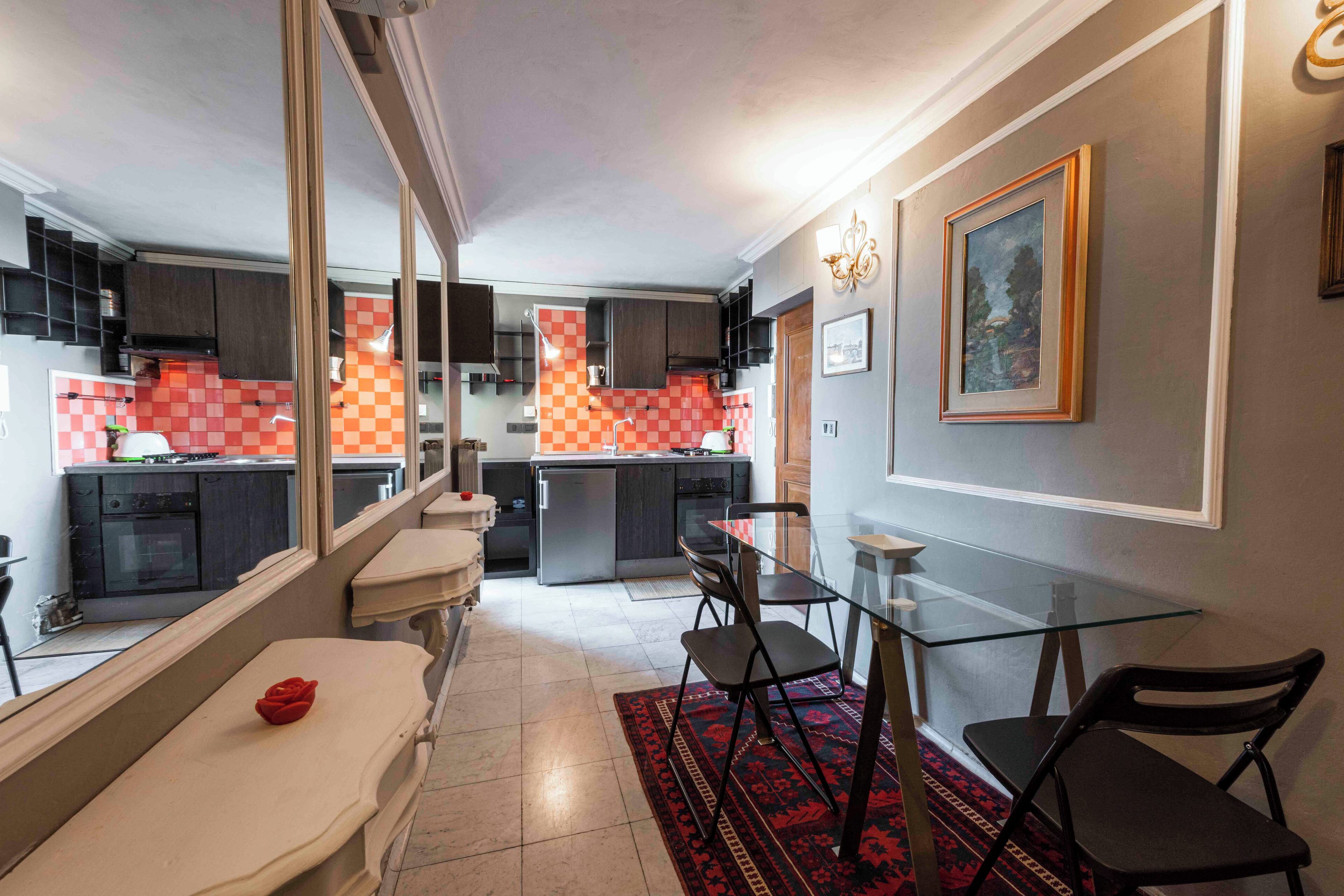 Een moderne keuken met een centraal eiland, donkere kasten en een zwart-wit geruit spatwand. Er is een zithoek met een glazen tafel en stoelen, en een rode roos op een bankje.