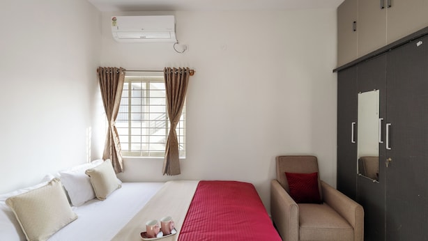 A hotel room with a bed, a chair, a wardrobe, and an air conditioning unit. - The Galleria Home by Red Olive Yelahanka (Bengaluru)