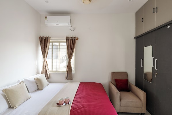 A hotel room with a bed, a chair, a wardrobe, and an air conditioning unit.